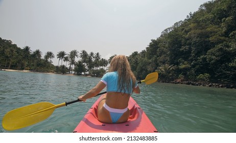 Pretty sexy young blonde woman in stylish swimsuit enjoys trip canoe on sea against palm islands. Traveling to tropical countries. Positive sports girl hand padding on kayak, back view.