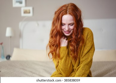 Pretty Shy Young Redhead Woman Sitting Relaxing On Her Bed Looking Down With A Gentle Smile, With Copyspace