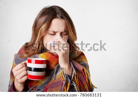 Pretty sick woman has runnning nose, rubs nose with handkerchief, drinks hot beverage, wrapped in warm blanket, has high temperature and cold, isolated over white background. Sneezing female