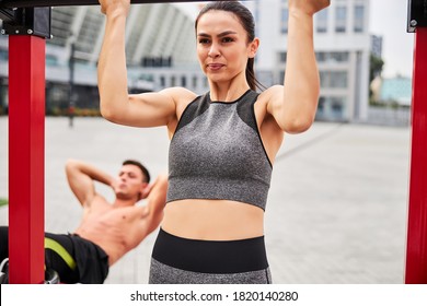 Pretty slim female is doing pull ups on horizontal bar while muscular male is doing abs crunches on sports ground - Powered by Shutterstock