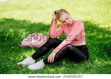Pretty slim fit blond woman in sportswear and sportsbag sitting and resting on the lawn in the park