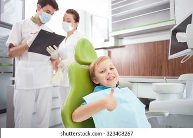 Pretty Small Boy Is Sitting In Medical Chair. He Is Giving Thumb Up And Smiling. The Dentist And Female Assistant Are Standing And Discussing With Seriousness. The Man Is Writing Documents