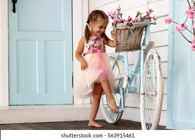 Pretty small girl in beautiful dress stay near the vintage white bicycle with basket with flowers over white wooden wall, beauty and fashion concept, outdoor portrait