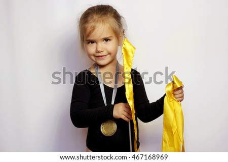 Pretty small girl in black sportswear with yellow ribbon with gold medal on her neck over white background, rhythmic gymnastics, indoor portrait, sport and health concept