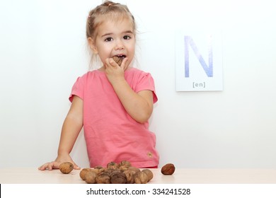 Pretty small girl cracking walnut at the desk with scattered nuts over white background with N letter on it, indoor portrait