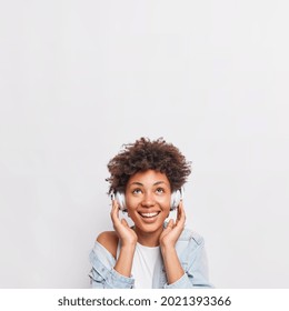 Pretty smiling African American woman listens music via headphones smiles broadly enjoys favorite playlist wears fashionable clothes poses against white background with blank space overhead. - Powered by Shutterstock