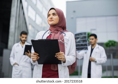 Pretty smiling Arab female doctor in hijab poses outdoors in front of modern clinic and looks away. Two confident male doctors standing behind and working with MRI scan and tablet pc - Powered by Shutterstock