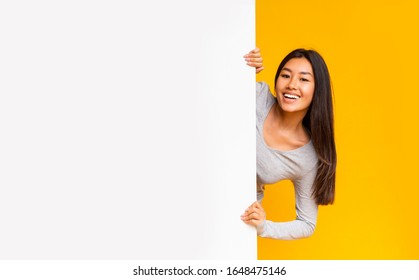 Pretty smiling asian girl looking from behind white advertising board over yellow background