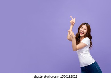 Pretty smiling Asian woman in casual white t-shirt pointing hand and looking to copy space aside on purple studio background