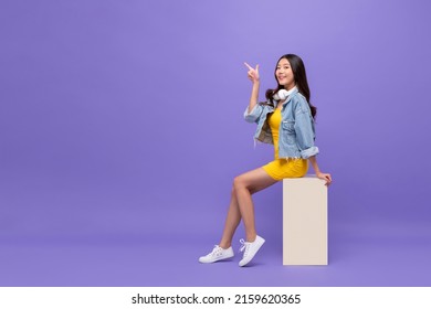Pretty smiling Asian woman sitting and pointing finger up in isolated purple color studio background with copy space - Shutterstock ID 2159620365