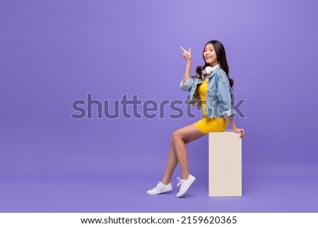 Pretty smiling Asian woman sitting and pointing finger up in isolated purple color studio background with copy space