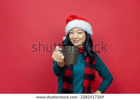 Pretty smiling asian woman in warm christmas sweater and santa hat is having hot chocolate on red background for season celebration