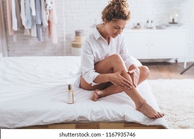 Pretty Smiling Caucasian Woman Applying A Lotion To Her Legs During Her Morning Routine.