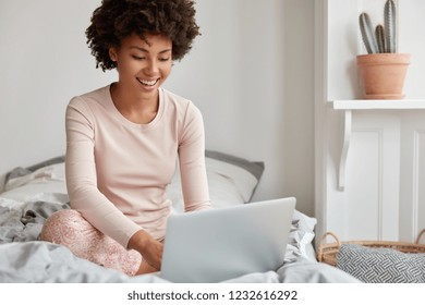 Pretty smiling ethnic girl has pleasant smile, Afro haircut, dressed in casual domestic clothes, laugh during watching comedy, leaves comments, chats online with friends on laptop computer sits in bed - Powered by Shutterstock