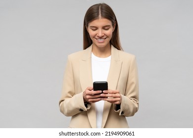 Pretty Smiling Female College Student In Beige Jacket Rehearsing Graduation Speech On Gray Copy Space Background Using Smartphone As Cheat Sheet. Education And Technology