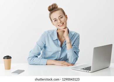 Pretty Smiling Female Freelance Remote Worker Wear Glasses Using Laptop Drinking Coffee Checking Emails Via Smartphone Working Leaning On Table In Office Smiling Happy. Gig Economy Concept