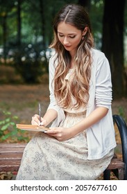 Pretty smiling girl sitting on bench in park and writing a letter. Outdoor portrait of beautiful young woman with pen and paper (notepad)