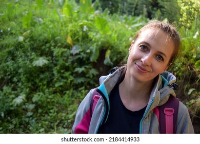 Pretty smiling hiker girl in anorak against the green grass background in the summer morning. Portrait of a cute backpacker lady after waking up in the morning in the summer mountains of Kazakhstan.