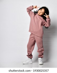 Pretty Smiling Little Curly-haired Asian Girl In Warm Pink Jumpsuit Standing And Showing Her Face With Her Fingers Against Her Eyes Against The Background Of A White Wall. Trendy Kids Fashion, Stylish