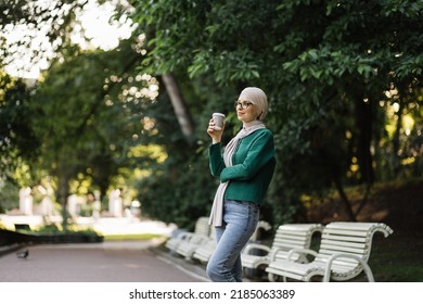 Pretty smiling muslim woman in hijab walks at green summer city park with coffee to go. Happy arabic girl standing outdoor with cup of hot drink. Lifestyle concept.