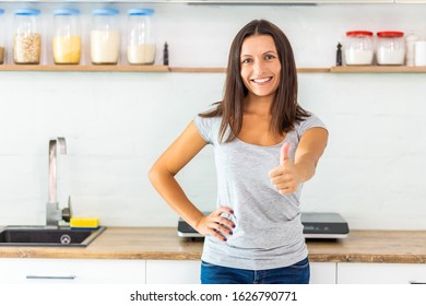 Pretty Smiling Woman With Dark Hair Looking At The Camera, Giving Thumb Up, Spending Time In Her New Beautiful Cozy Kitchen.