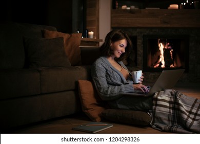 Pretty smiling woman drinking tea by the fireplace and looking at laptop at night. - Powered by Shutterstock