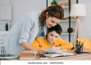 pretty smiling woman helping adorable son doing schoolwork at home
