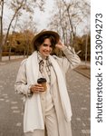 pretty smiling woman walking in park in autumn clothes, wearing beige trench, hat and white knitted scarf, winter cold season accessories trend, stylish outfit, elegant beautiful lady with coffee