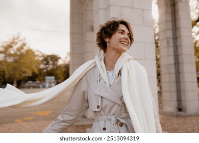 pretty smiling woman walking in park in winter clothes, portrait in beige trench and white knitted scarf, winter cold season accessories trend, stylish outfit, elegant beautiful lady, short haircut - Powered by Shutterstock