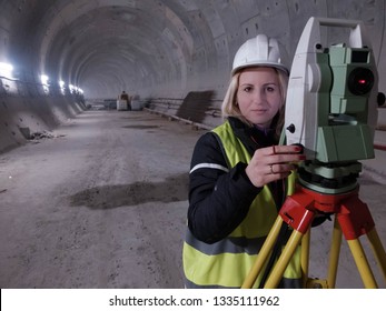 Pretty smiling young mining surveyor woman stands by geodetic total station in the tunnell of metro under construction