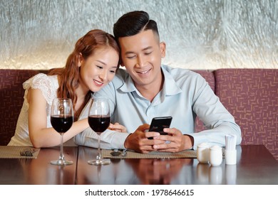Pretty smiling young woman leaning on shoulder of boyfriend and watching funny video on smartphone when waiting for food in restaurant