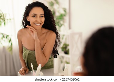 Pretty Smiling Young Woman Touching Her Face With Silky Smooth Skin Near Mirror At Home, Empty Space. Beautiful Millennial Female Pampering Herself After Bath, Wearing Towel