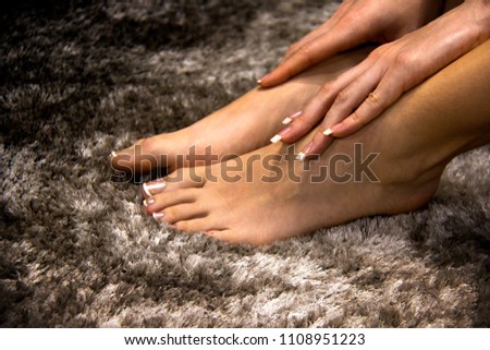 Pretty and soft woman feet touched by hands close up, white and pink transparent french manicure on her nails and toes, side view on grey background