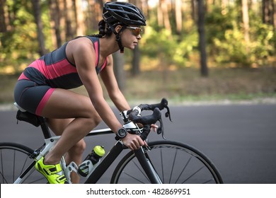 Pretty sportive girl rides a bike on the road on the nature background. She wears black-pink sportswear, a stopwatch, a black helmet, sunglasses and green sneakers. Shoot from the side. Horizontal.