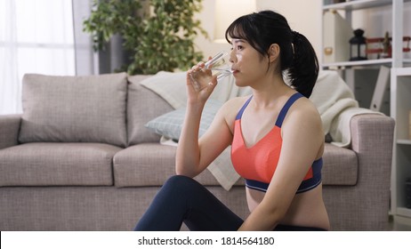 Pretty Sporty Korean Woman Letting Out A Small Sigh Of Relief With Contented Look After Taking A Sip Of Water And Relaxing By Couch.