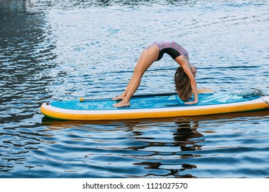 Pretty, sporty woman in bridge position with straight legs on paddleboard, doing yoga on sup board at sea