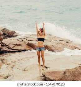 Pretty sporty woman in jeans short shorts, red headband and black bra stand on big stone on the beach during sea ocean storm. Big waves behind her. Concept of danger, sad, bad emotions.