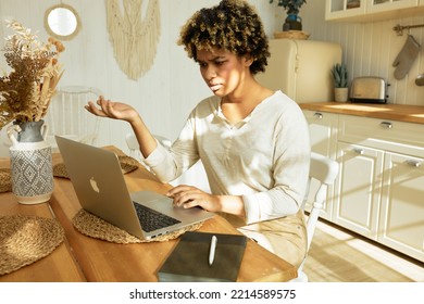 Pretty Student Girl Sitting At Kitchen Table In Boho Style Interior Studying Online On Laptop, Gesticulating With Hand And Uncertain Face Expression Trying Figure Out How To Use Learning Application