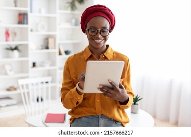 Pretty Stylish African American Woman In Casual Outfit Standing By Worktable, Using Digital Tablet And Smiling, Posing Alone At Cozy Office, Copy Space. Gadgets And Business Concept