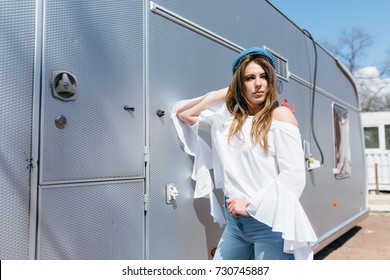 Pretty Stylish Girl With Long Dark Hair And Charming Smile Dressed In White Shirt And Fashion Cap. She Is Posing To Camera In The Park On The Background Trailer. Beautiful Girl On The Street