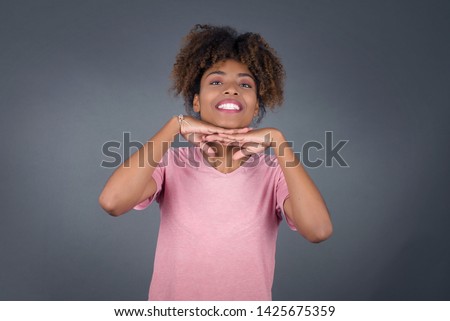 Pretty surprised African American lady in white pants fooling around in studio and jumping. Adorable girl in knitted sweater dancing on colorful background and touching chin with hands