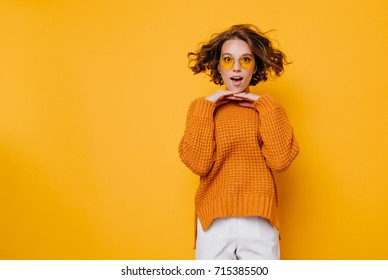 Pretty surprised lady in white pants fooling around in studio and jumping. Adorable girl in knitted sweater dancing on colorful background and touching chin with hands.