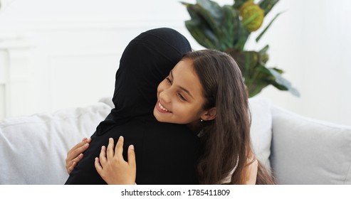 Pretty Teen Small Daughter Talking With Muslim Mother In Hijab And Embracing. Arab Little Girl Having Talk With Woman In Headscarf On Couch At Home And Hugging. Mom's Love Hugs. Rear. Back View.