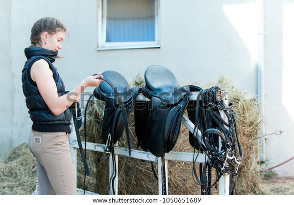 Pretty teenage girl equestrian cleans\
black Leather Horse Saddle and equipment at farm on bright sunny\
day. Horizontal outdoors summertime\
image.