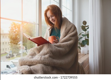 pretty thoughtful smiling young girl reading book and drinking morning coffee at home sitting next to the window wrapped in warm comfy blanket