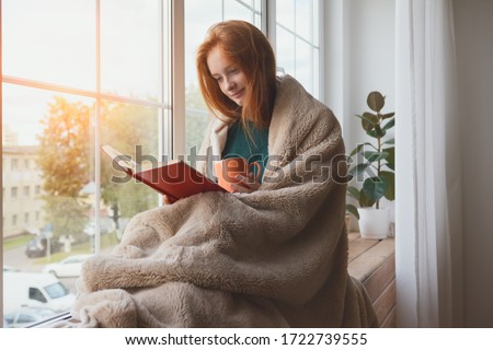 pretty thoughtful smiling young girl reading book and drinking morning coffee at home sitting next to the window wrapped in warm comfy blanket