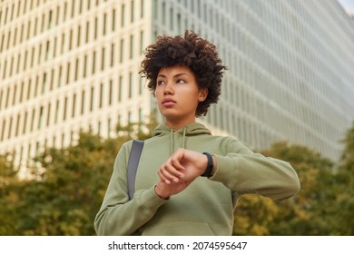Pretty thoughtful sportswoman waits for trainer outdoors checks time on wristwatch dressed in sweatshirt poses against modern city building outdside does regular exercising. Healthy lifestyle