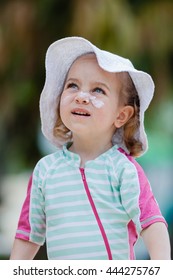 Pretty Toddler Girl In Spf Solar Suit, Hat And With Sunscreen Cream On Her Face Playing At The Beach. Safety In Summer Sun.