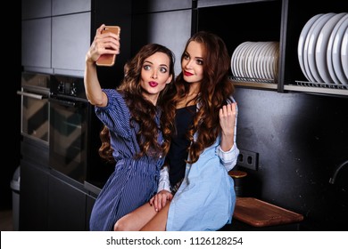 Pretty twins young women together in a kitchen room making photo on mobile phone, casual home style