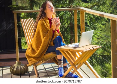 Pretty white girl is sitting relaxing on balcony wrapped in yellow plaid holding cup of tea, enjoying coffee. Young brunette woman works online, remotely on laptop. Sweet home in a wood at autumn day.